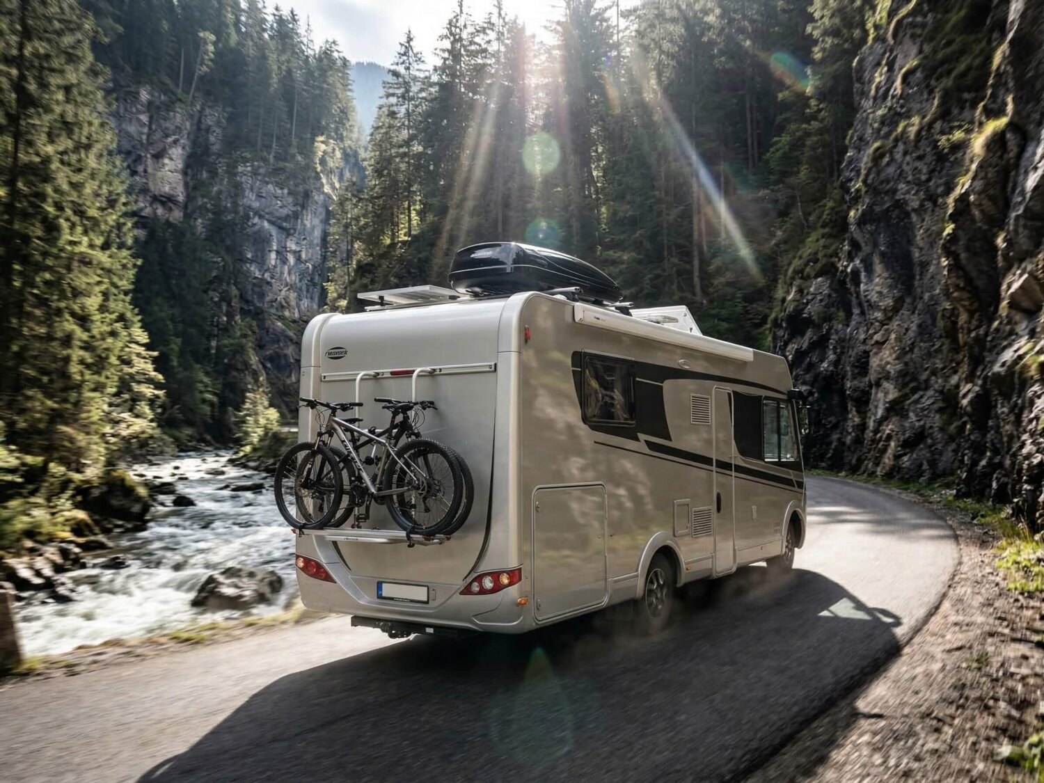 Autocaravana en carretera de montaña junto a un río, con bicicletas y bosque de coníferas.