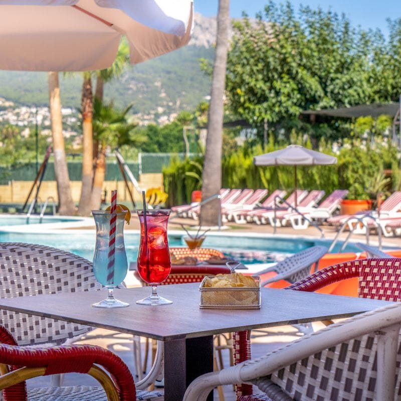 Mesa exterior junto a piscina con dos cócteles coloridos y aperitivos en zona de relax del camping con tumbonas y sombrillas al fondo