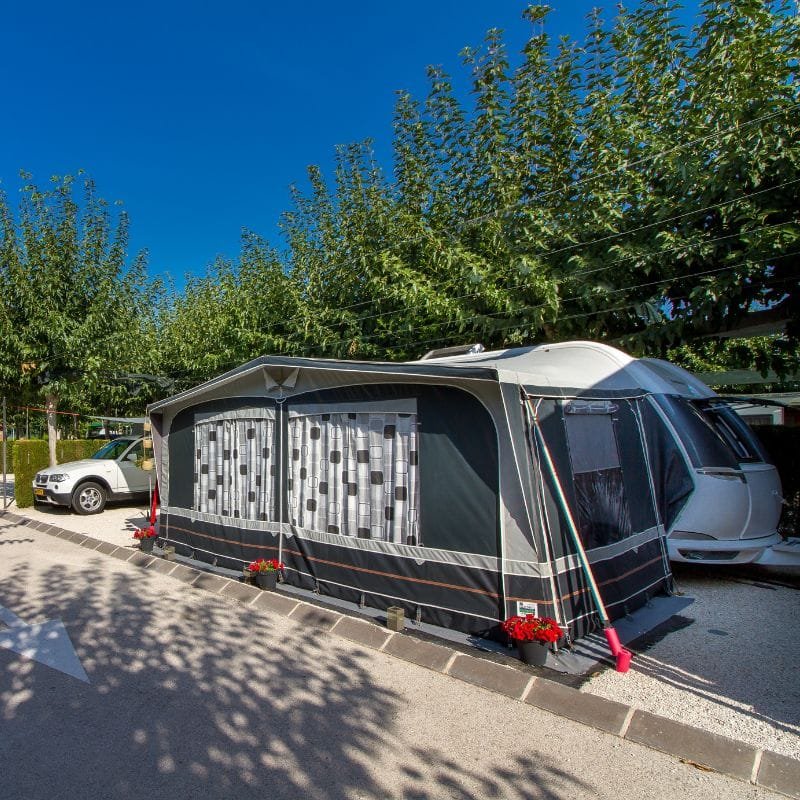 Avance o tienda exterior montada junto a una caravana en parcela de camping con plantas decorativas y sombra de árboles
