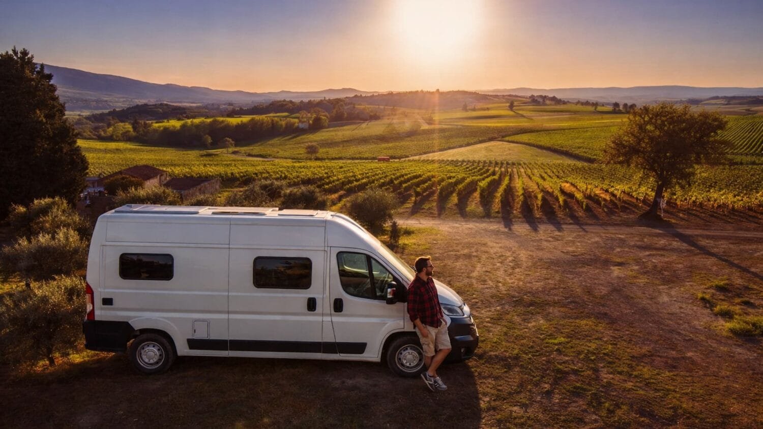 Furgoneta camper blanca estacionada en paisaje rural con viñedos al atardecer y viajero disfrutando del entorno