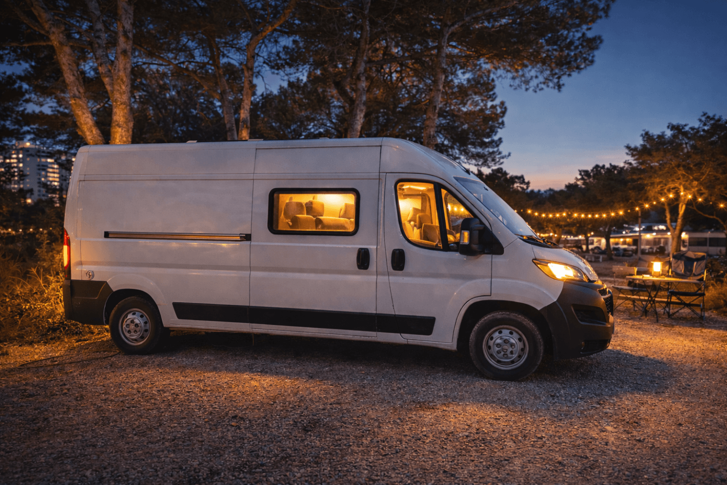 Camper blanca estacionada en camping por la noche con luces cálidas y zona de descanso exterior iluminada