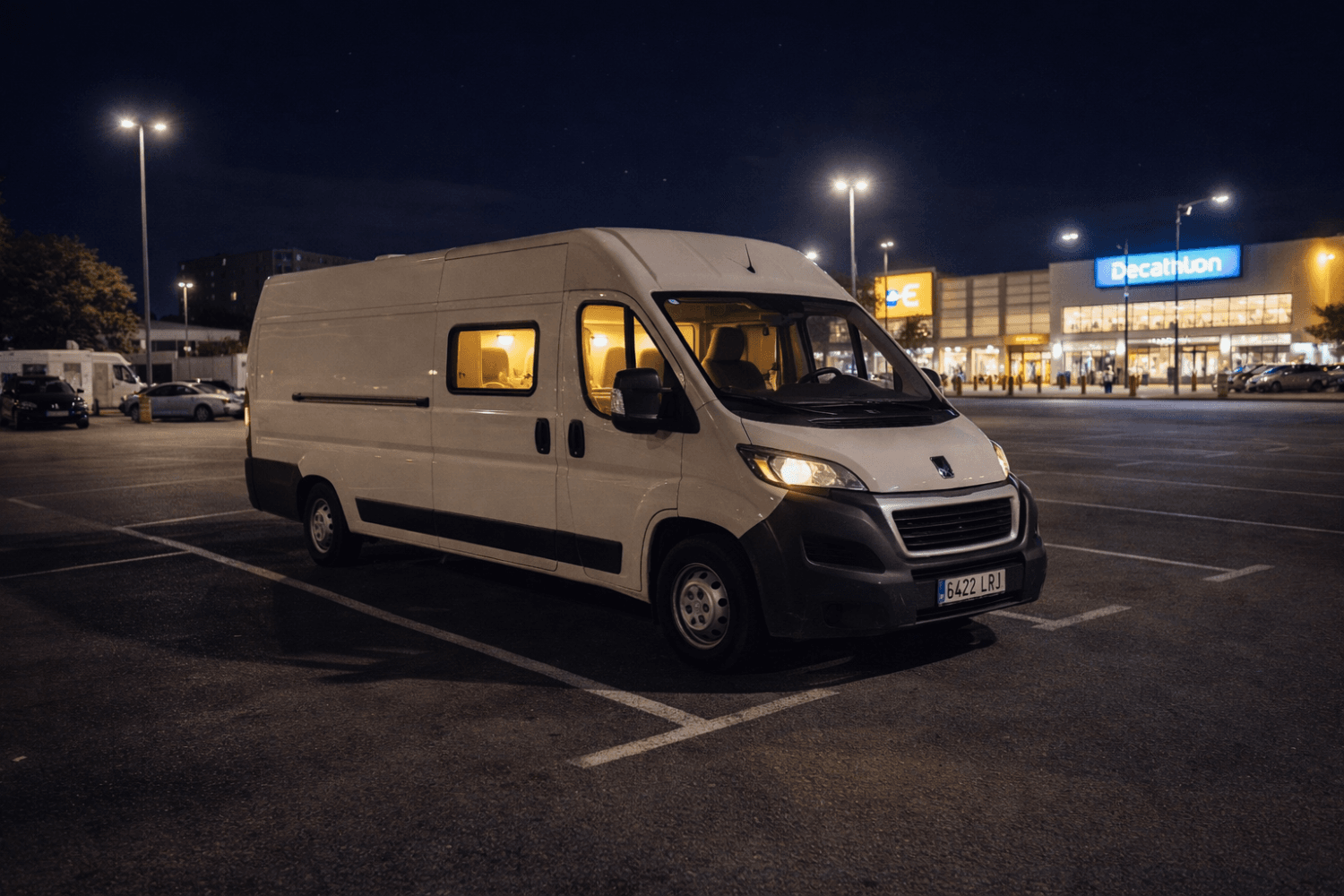 Camper blanca aparcada de noche en parking iluminado frente a centro comercial