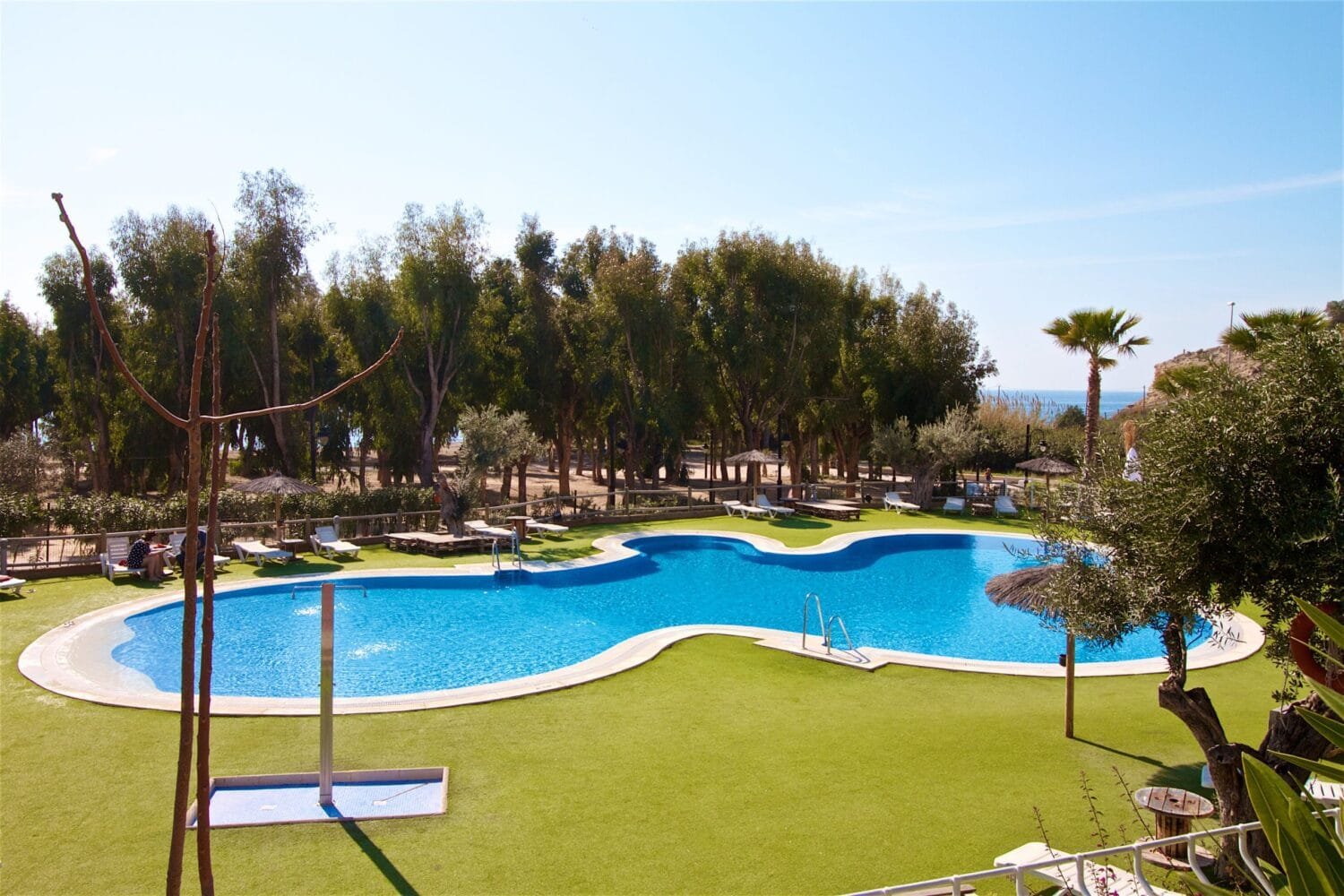 Piscina exterior de camping con forma irregular rodeada de césped, tumbonas y zona arbolada junto al mar