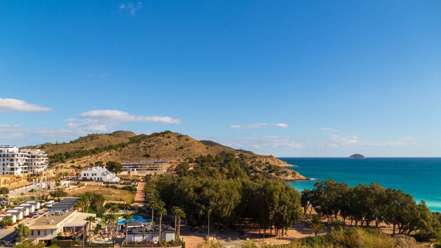 Vista panorámica de costa mediterránea con playa, colinas, urbanización y mar azul en día despejado