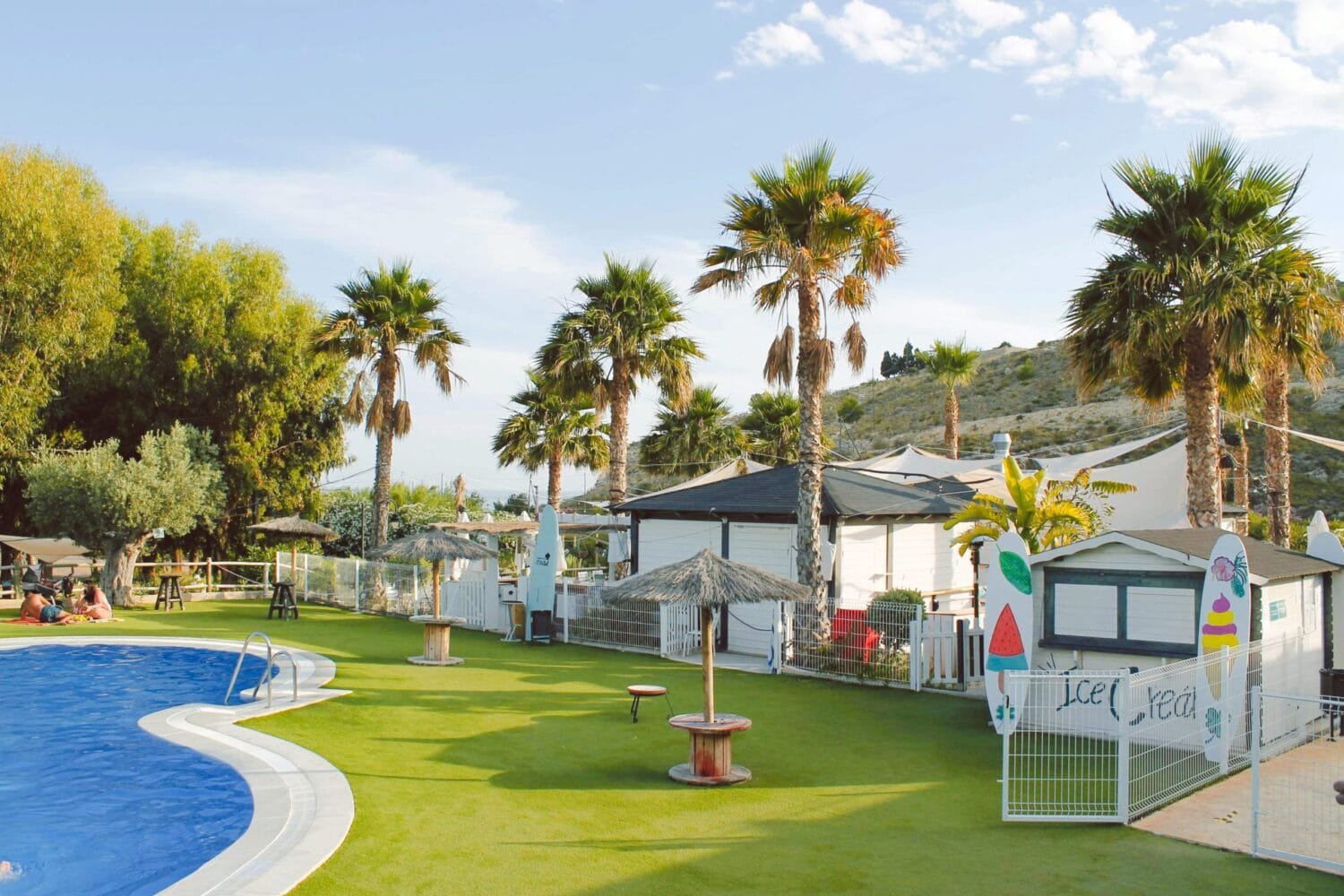 Piscina de camping rodeada de palmeras, césped artificial y bungalows con zona infantil y sombrillas de paja