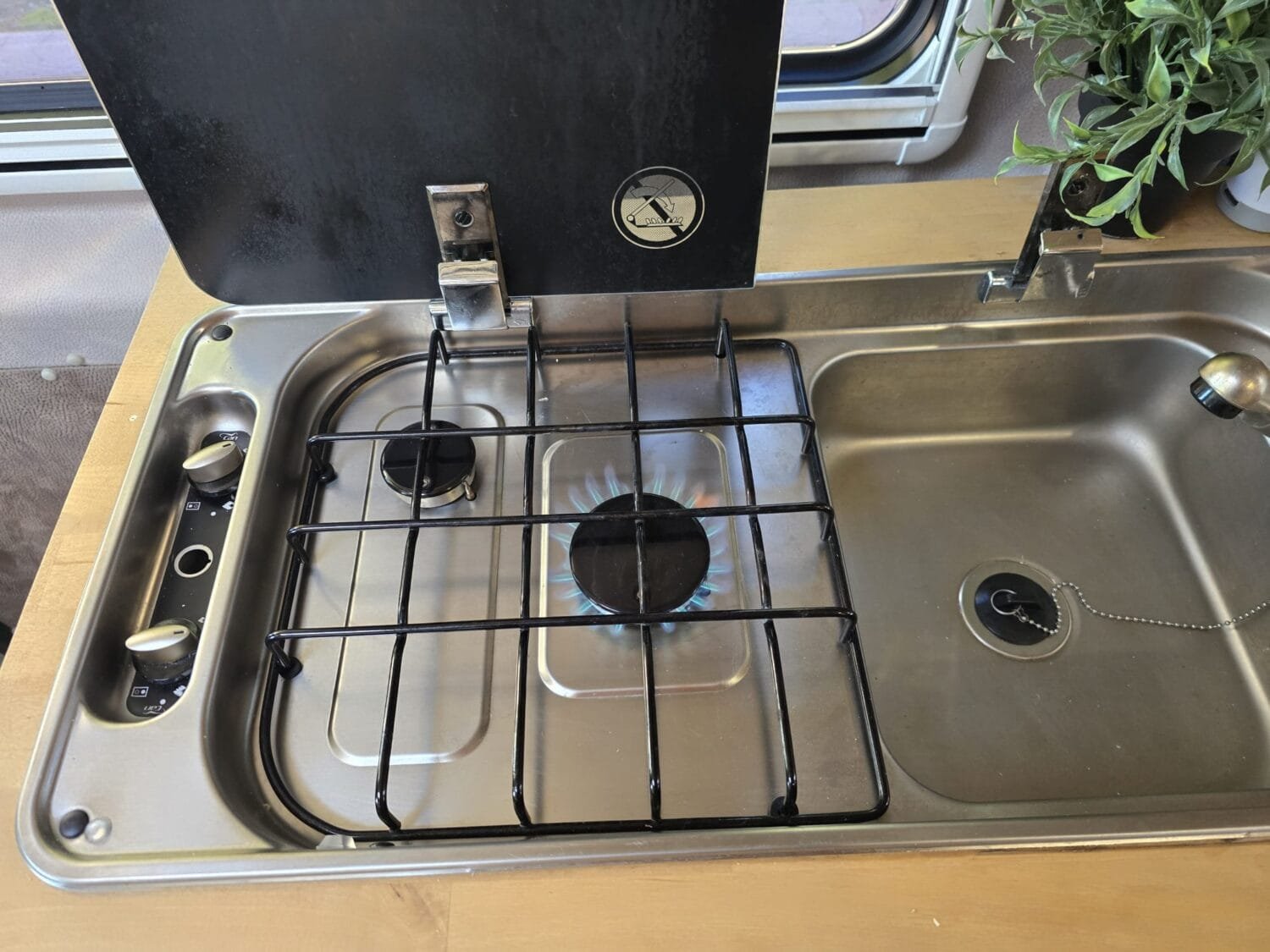 Cocina de gas de dos fuegos integrada junto al fregadero de acero inoxidable en la autocaravana, con tapa abierta y un quemador encendido