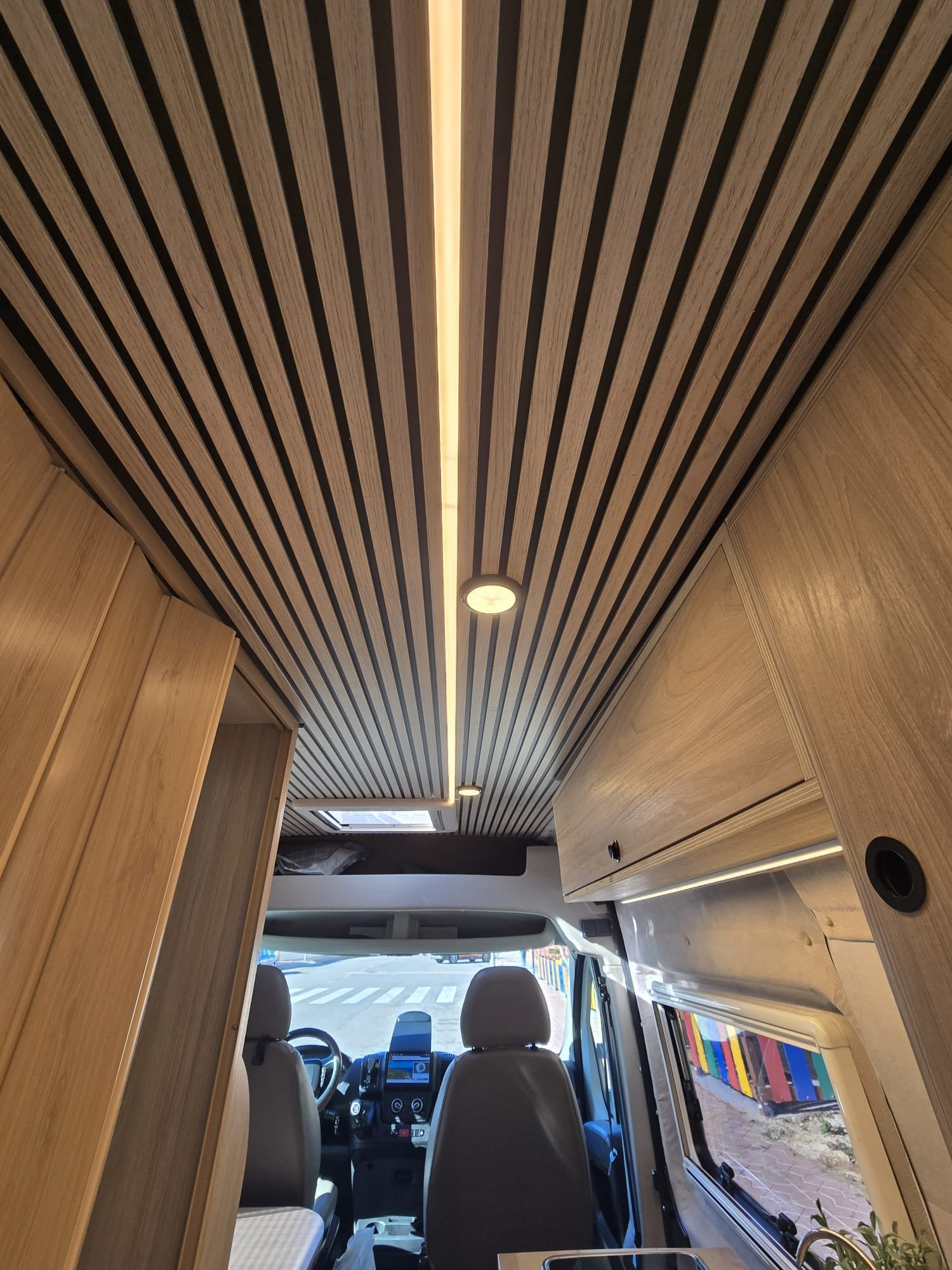 Techo interior de autocaravana con paneles de madera, iluminación LED lineal y focos empotrados sobre armarios superiores y cabina delantera