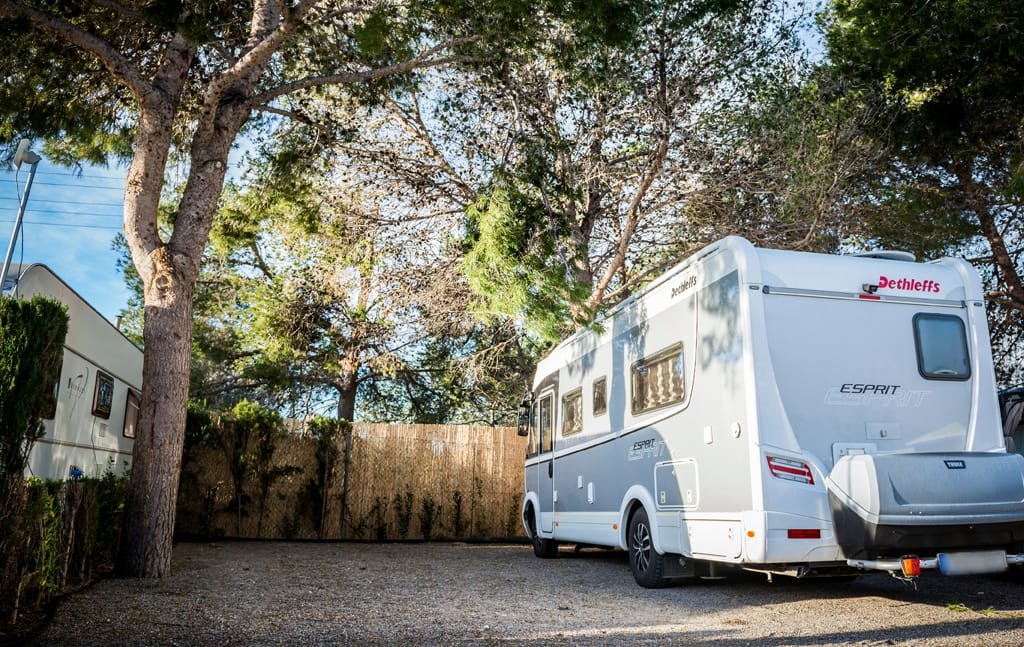 Parcela de camping sombreada con autocaravana estacionada bajo pinos y espacio delimitado para acampada en Benidorm