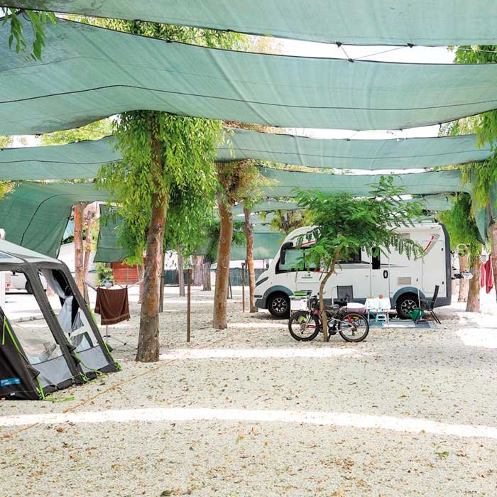 Parcela de camping con sombra natural y toldos verdes, autocaravana estacionada entre árboles y zona de acampada equipada