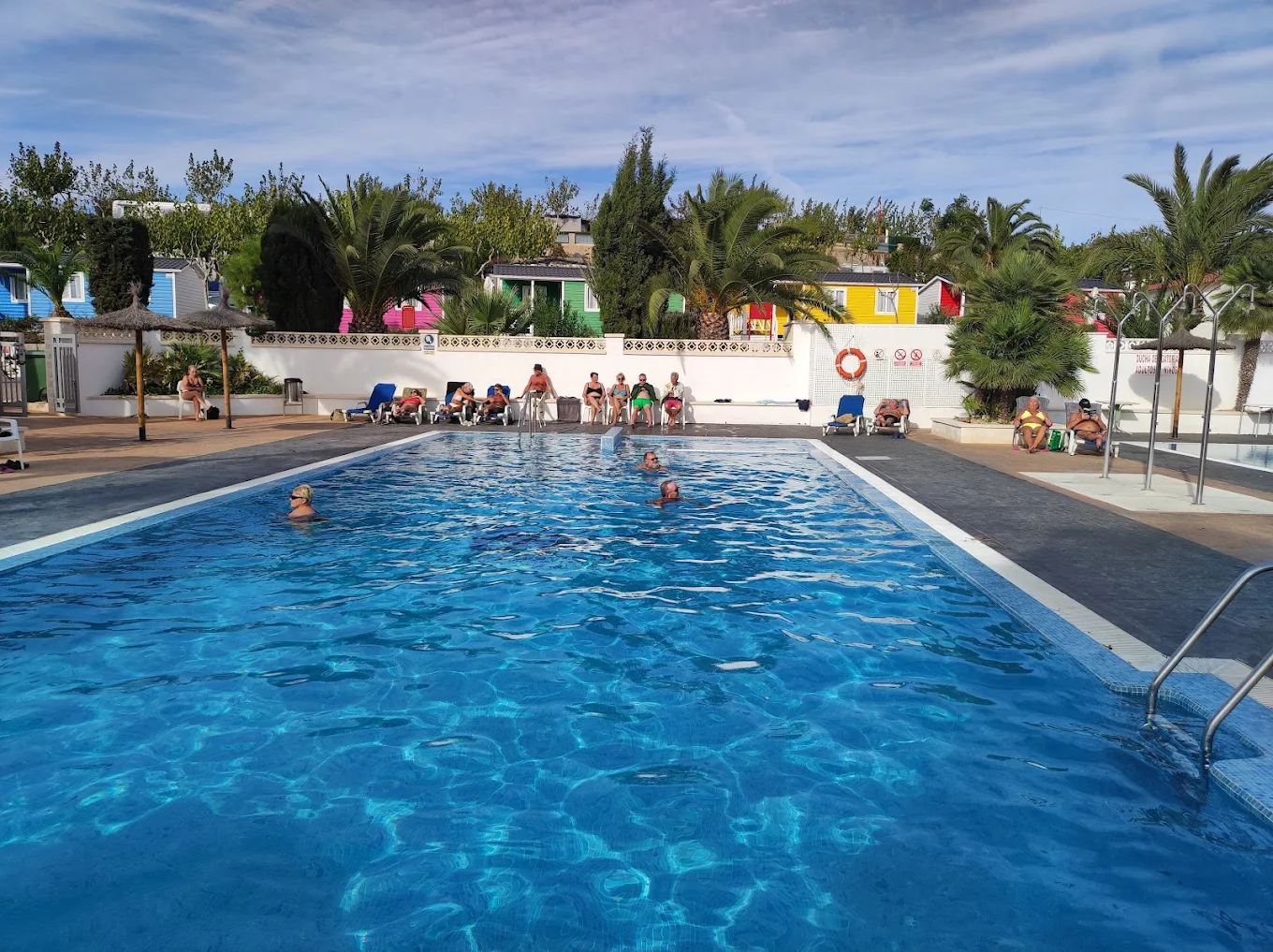 Piscina del camping con personas bañándose y zona de descanso con palmeras y bungalows al fondo
